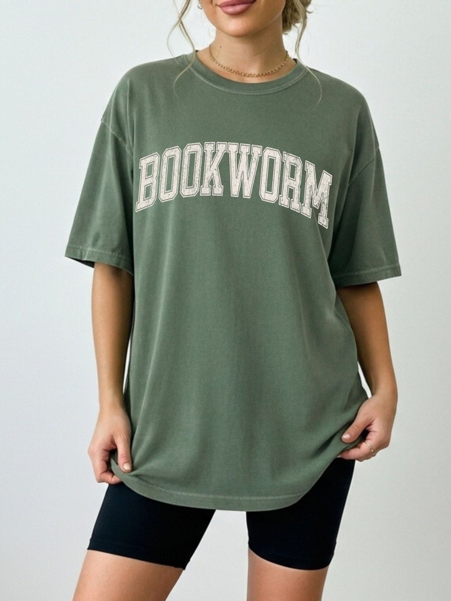 woman standing wearing a moss green comfort colors shirt that says "bookworm" in a varsity college style font