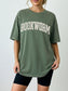 woman standing wearing a moss green comfort colors shirt that says "bookworm" in a varsity college style font