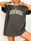 woman standing wearing a pepper grey comfort colors shirt that says "bookworm" in a varsity college style font