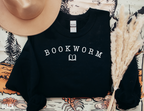 black bookworm sweatshirt folded with white varsity-style text and book graphic, styled with jeans, coffee, and a tan hat