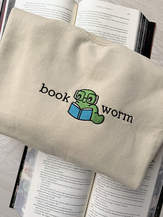 Embroidered bookworm design on sand-colored sweatshirt with a green worm and blue book, laid on open Rebecca Yarros novel, perfect for cozy book lovers

