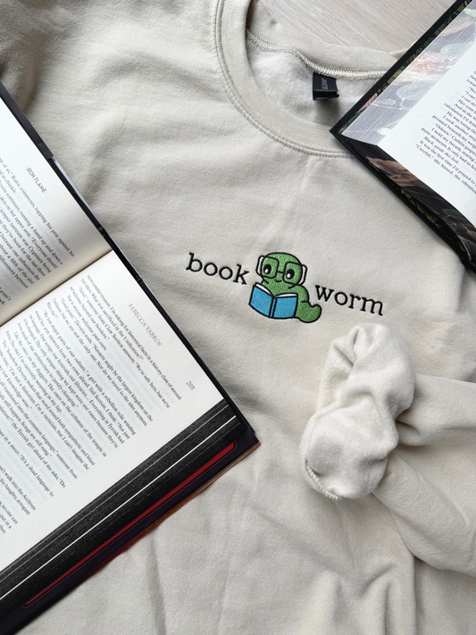 Sand sweatshirt featuring embroidered green worm reading a blue book with “bookworm” text, styled flat on top of Iron Flame pages 

