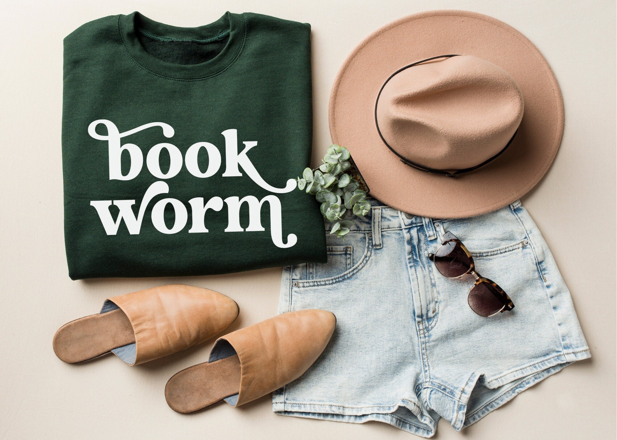 Folded green bookworm sweatshirt with white text next to tan hat, light denim shorts, sunglasses, and tan flats