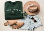 forest green bookworm sweatshirt folded with white text and book icon, styled with tan hat, sandals, and denim shorts