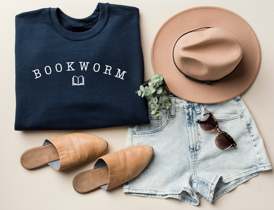 folded navy blue bookworm sweatshirt with white text and book graphic, styled with tan hat, sunglasses, and denim shorts