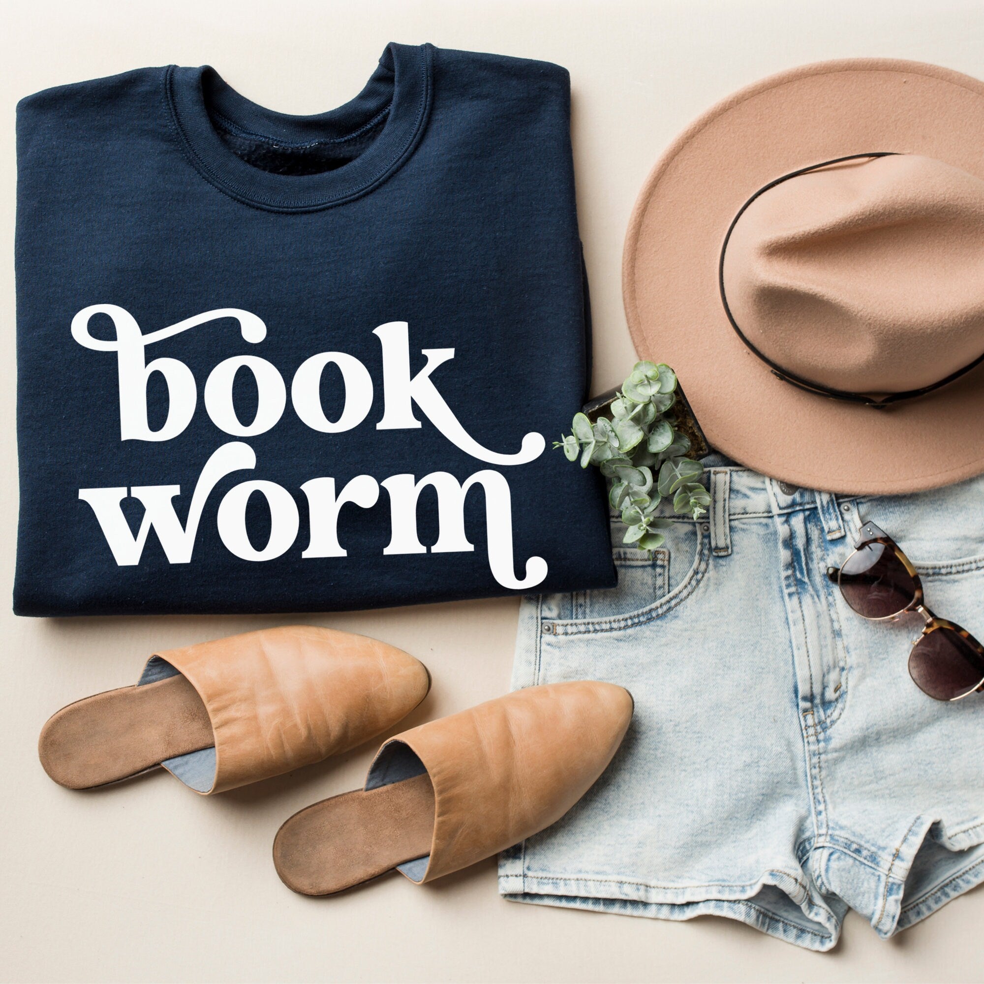 Folded navy sweatshirt with large white “bookworm” text, styled next to a tan hat, sunglasses, and light denim shorts