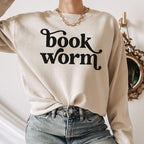Woman in sand-colored sweatshirt with bold black “bookworm” text in a playful serif font, accessorized with gold chain and jeans