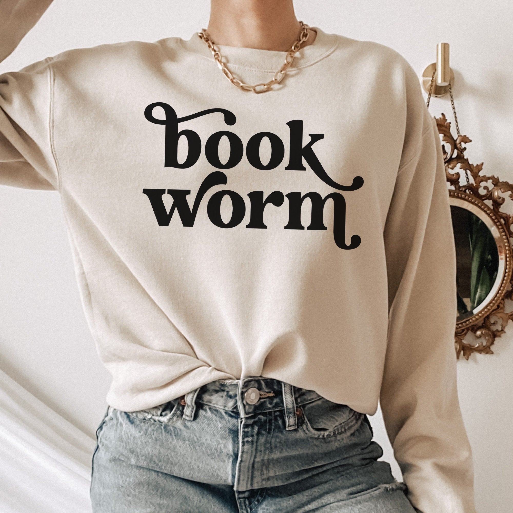 Woman in sand-colored sweatshirt with bold black “bookworm” text in a playful serif font, accessorized with gold chain and jeans