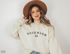 woman wearing a sand-colored bookworm sweatshirt with curved black text and open book icon, styled with gold chain necklace and denim jeans