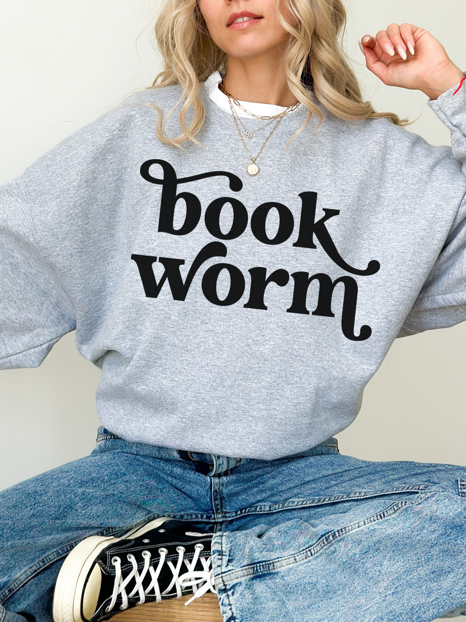 Woman in sport gray bookworm sweatshirt with large black text, sitting cross-legged in jeans and Converse sneakers