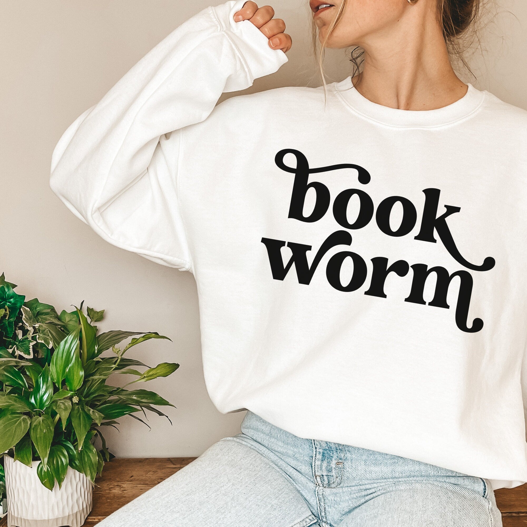 Woman in white sweatshirt with bold black “bookworm” text, sitting casually in a cozy space with plants