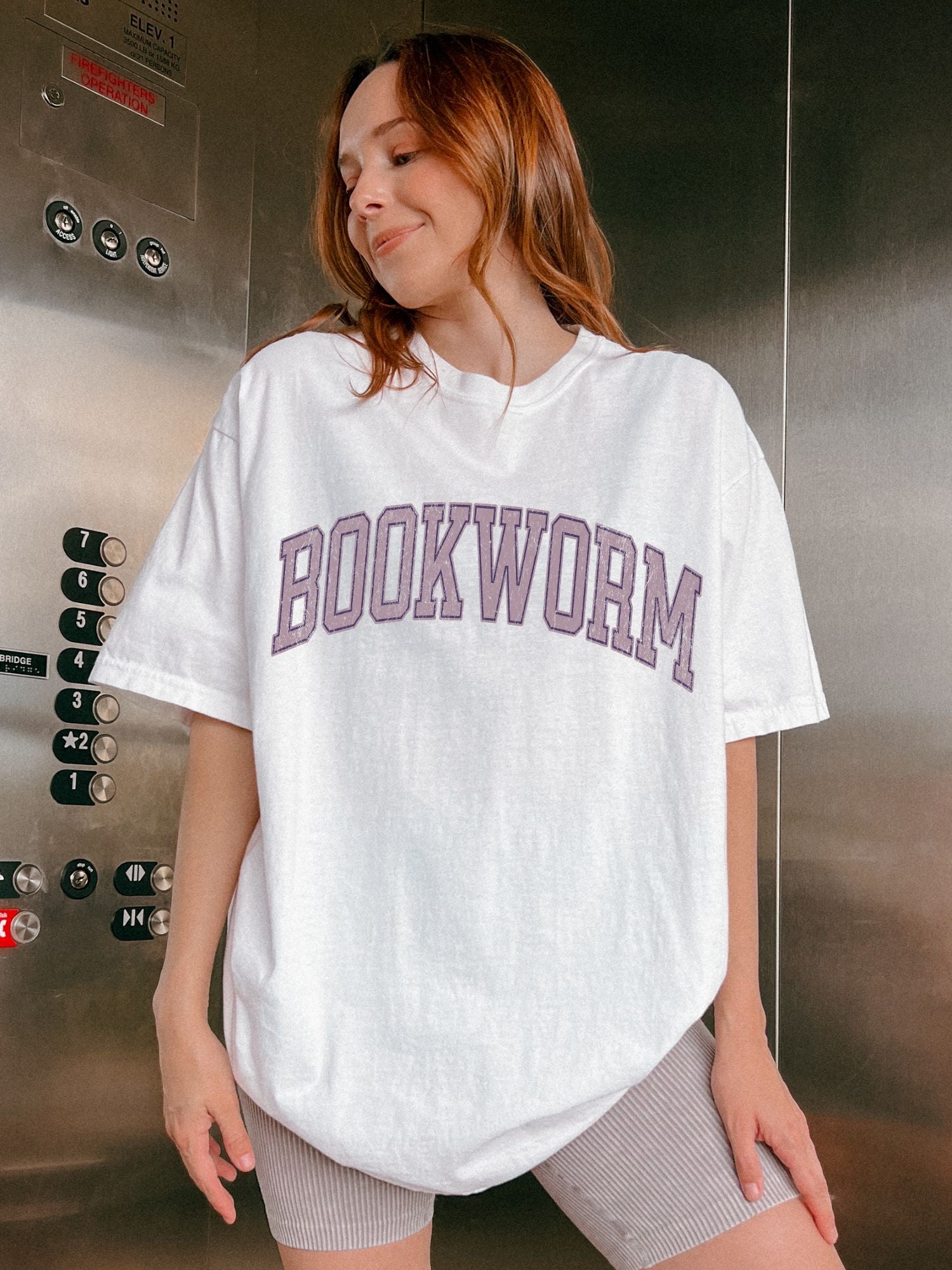 woman wearing a white oversized bookworm shirt with purple varsity lettering in elevator setting