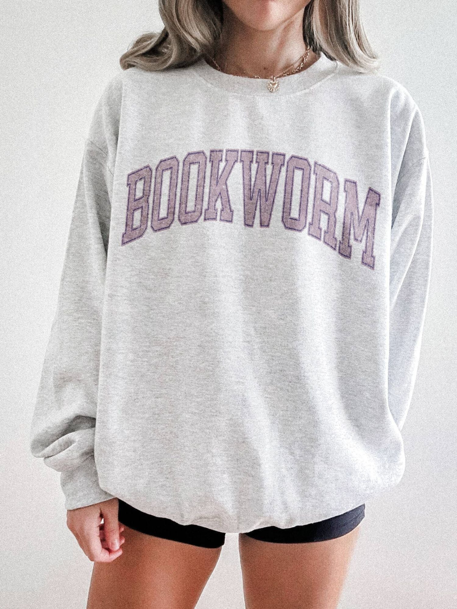 girl standing wearing an ash grey sweatshirt that says "bookworm" across the top in a jersey sport font