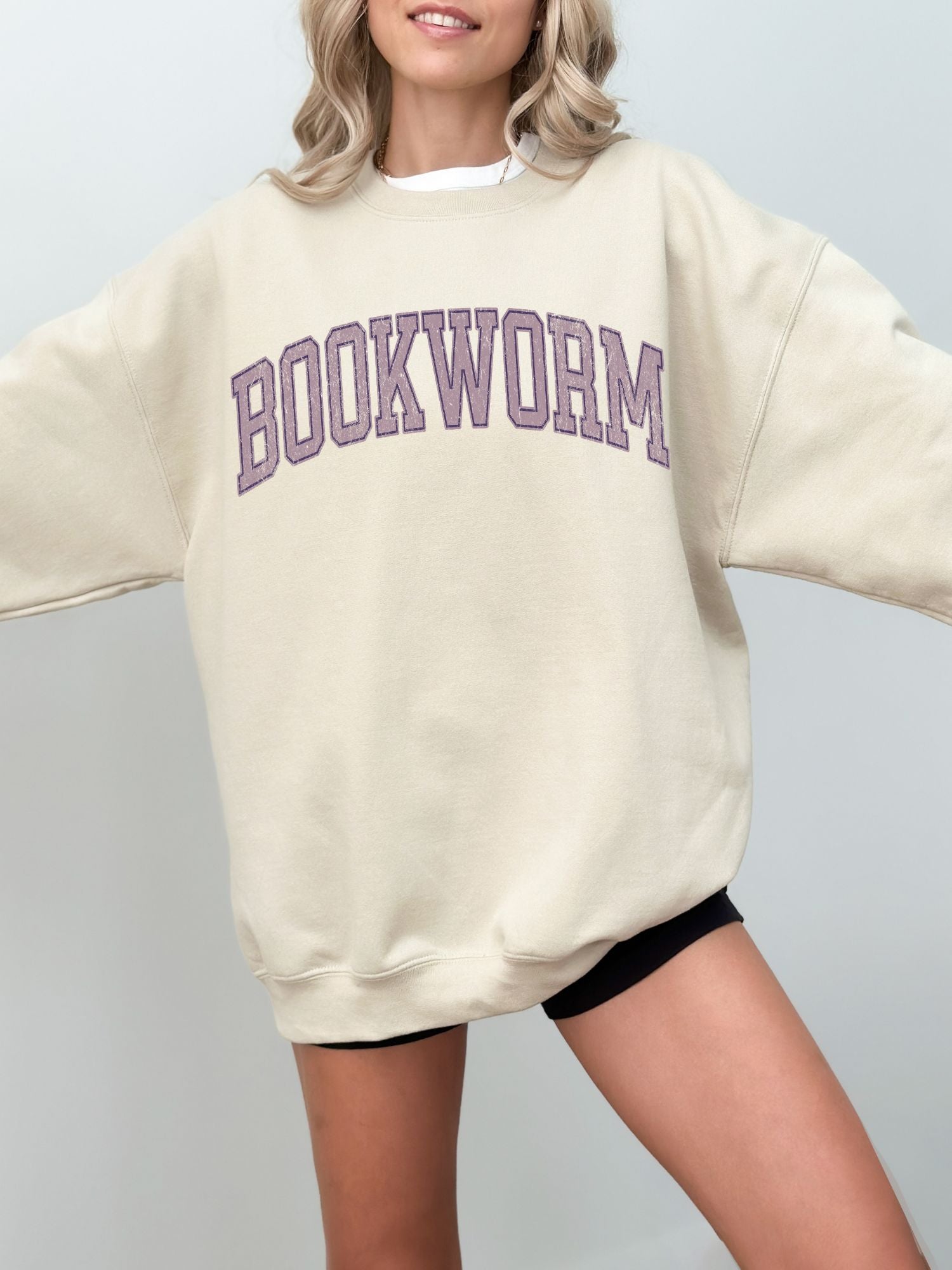 girl standing wearing a sand sweatshirt that says "bookworm" across the top in a purple jersey sport font