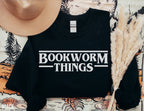 folded black sweatshirt with white bookworm things design laid out with pampas grass and hat