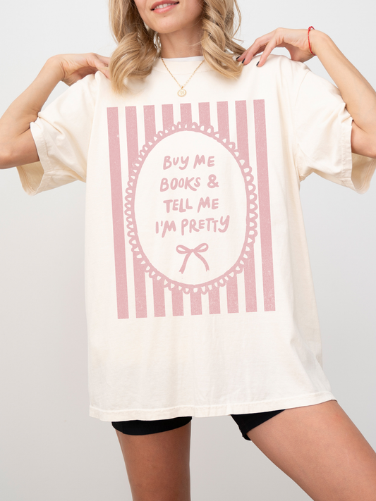 Woman wearing a white oversized t shirt with pink striped bow frame design and text buy me books and tell me I’m pretty standing in an elevator