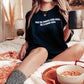 woman sitting on bed wearing black shirt that says buy me books and call me a good girl holding a coffee mug
