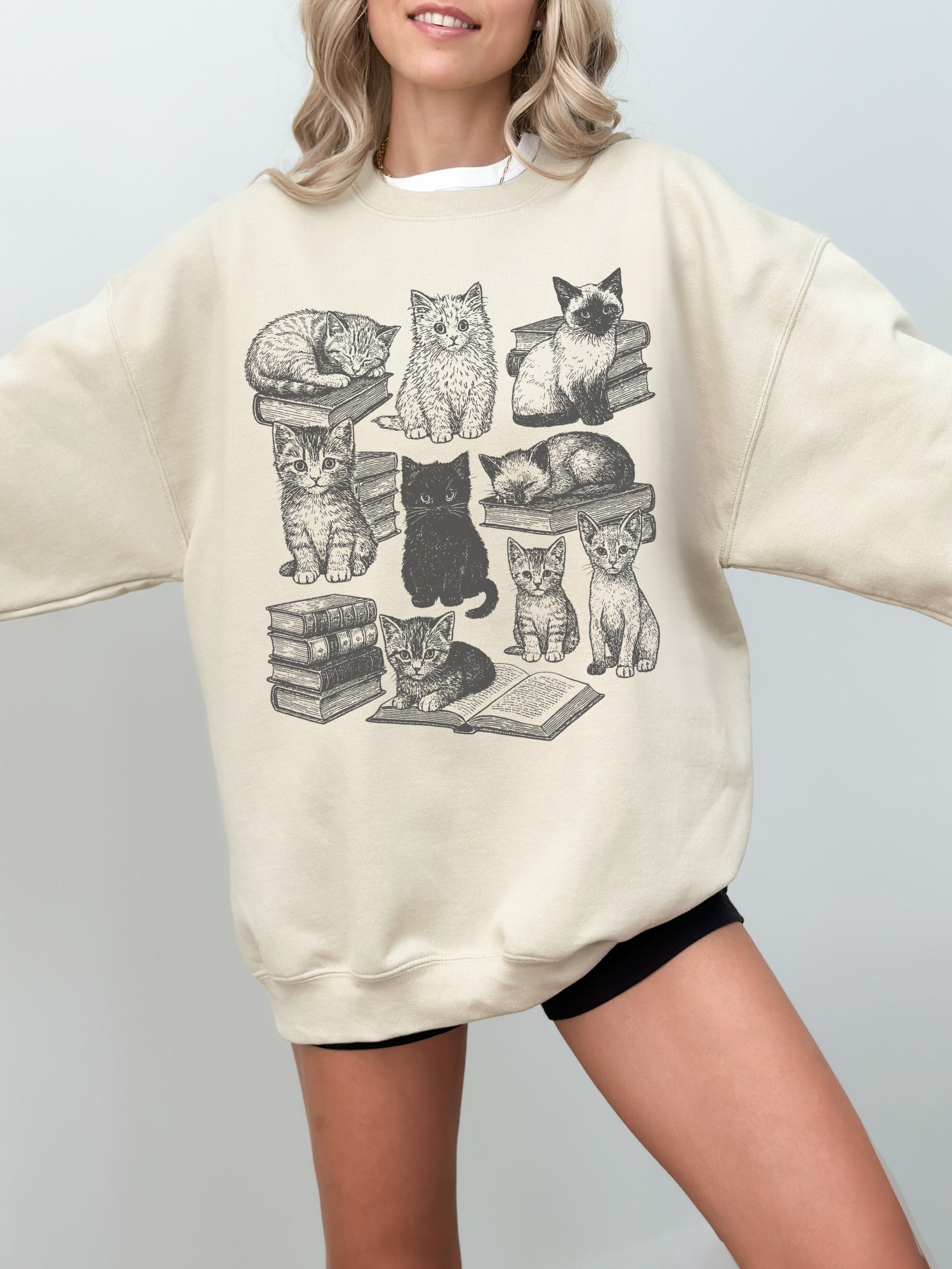 neutral beige sweatshirt showcasing hand-drawn cats on book piles and an open book