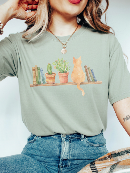 woman wearing a Bay green comfort colors t shirt with watercolor cat sitting on a shelf with books and potted plants