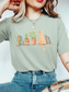 woman wearing a Bay green comfort colors t shirt with watercolor cat sitting on a shelf with books and potted plants