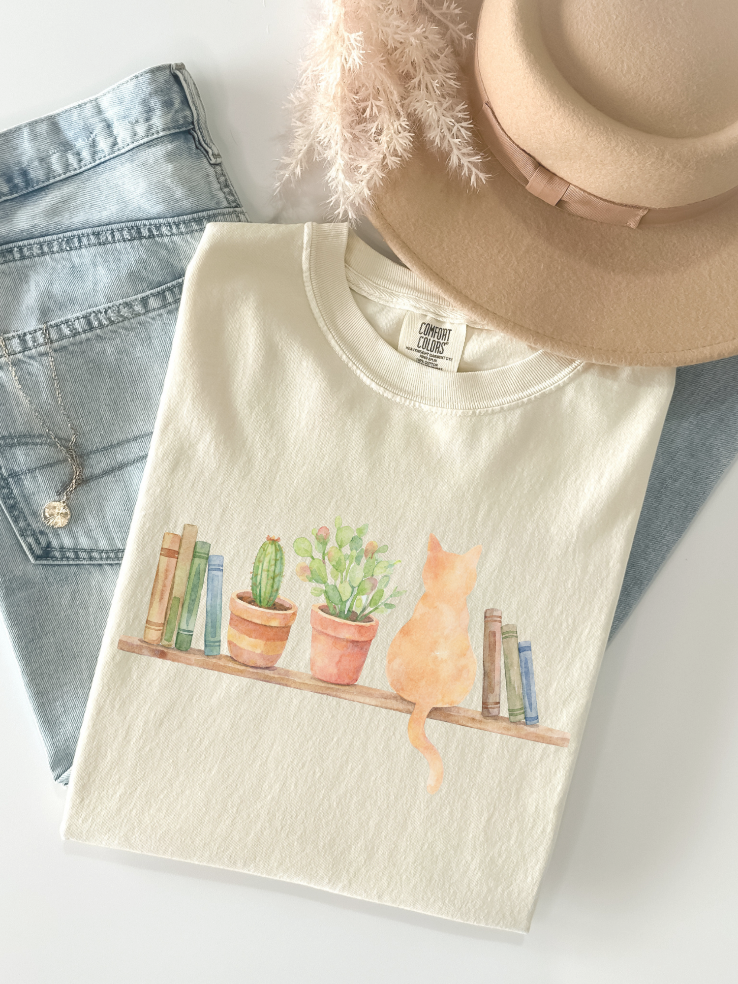 flat lay of Ivory comfort colors t shirt showing watercolor cat books and plants design styled with denim and hat