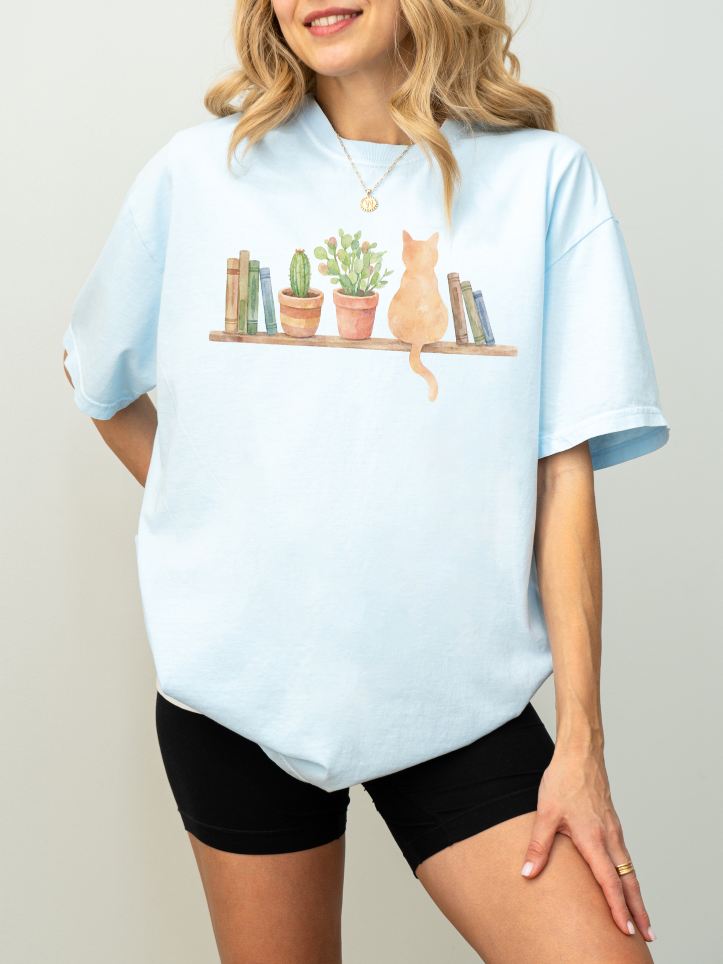 woman wearing a light blue comfort colors t shirt with a painted bookshelf scene featuring plants and a cat