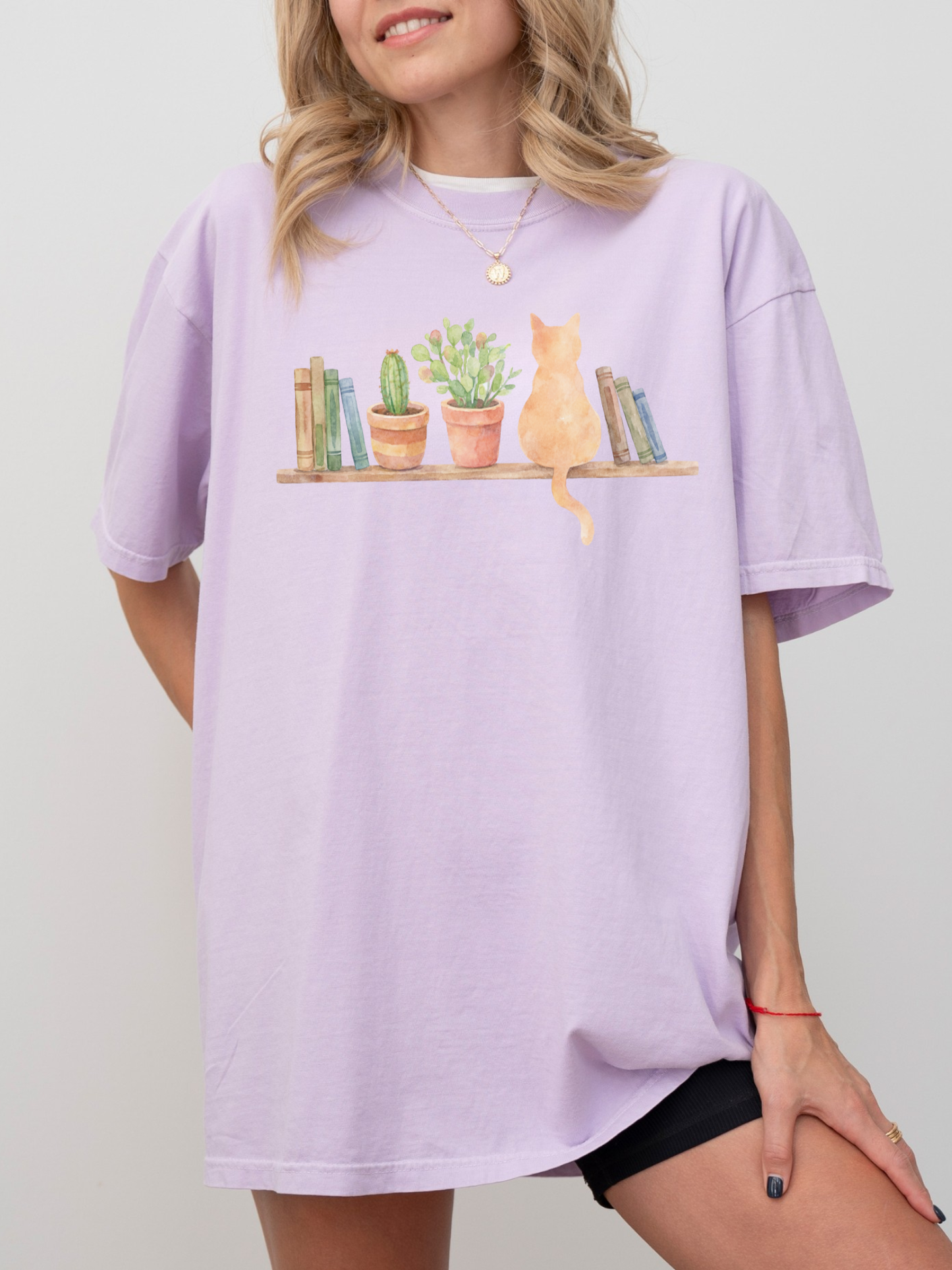 woman wearing a lavender comfort colors t shirt with watercolor cat books and houseplants on a wooden shelf