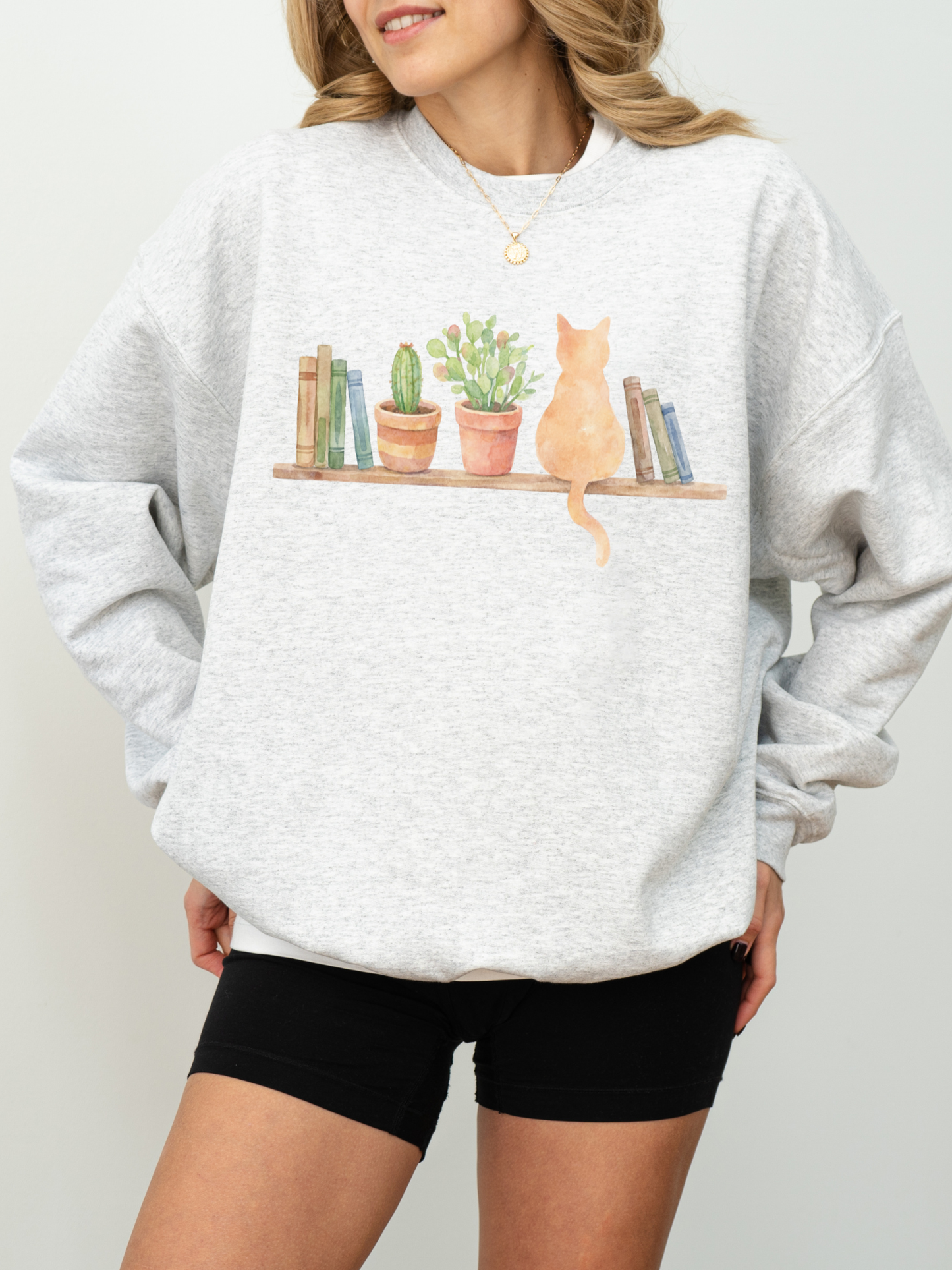 woman wearing an ash grey sweatshirt with watercolor cat sitting on a shelf with books and potted plants