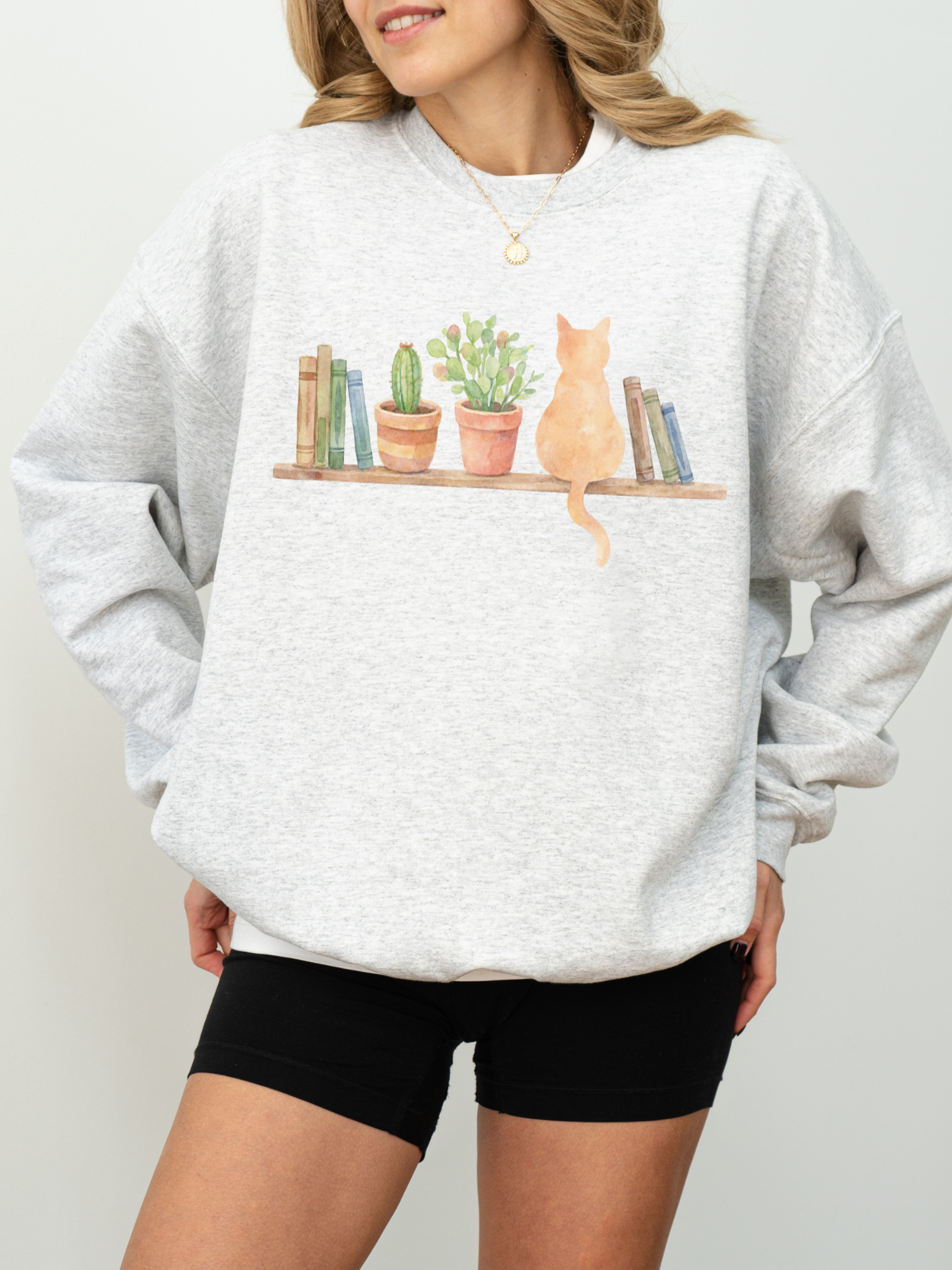 woman wearing an ash grey sweatshirt with watercolor cat sitting on a shelf with books and potted plants