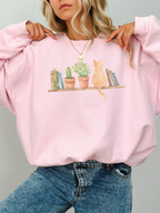 woman wearing a light pink sweatshirt with watercolor cat books and houseplants illustration