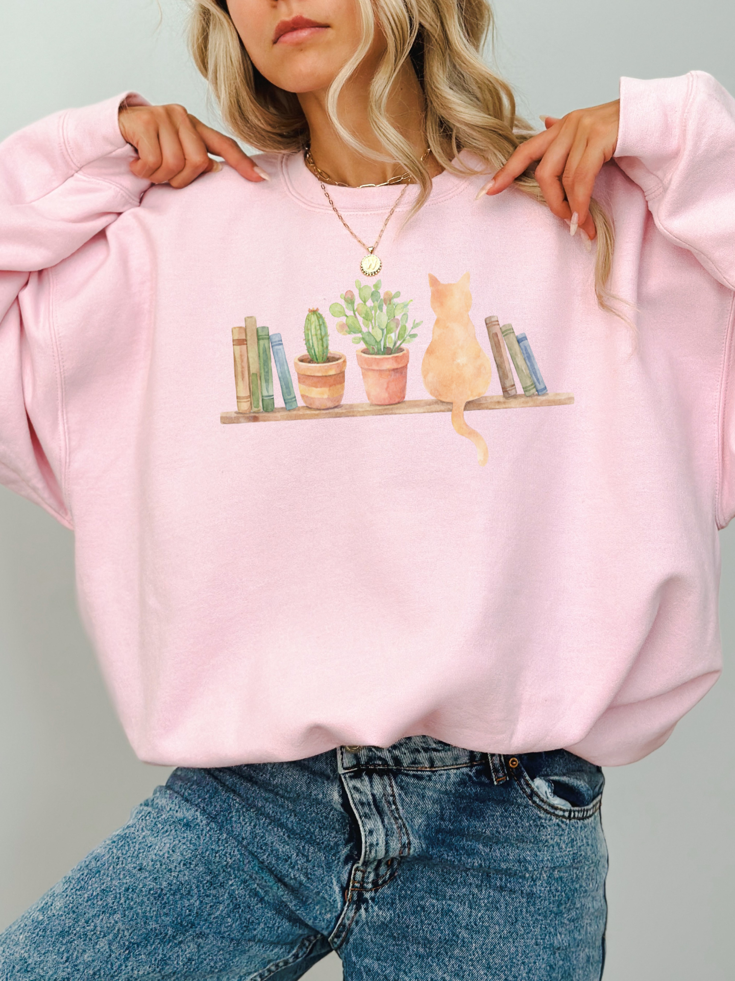 woman wearing a light pink sweatshirt with watercolor cat books and houseplants illustration