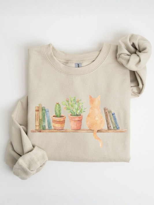 folded sand colored sweatshirt showing watercolor cat books and plants design on a flat lay background