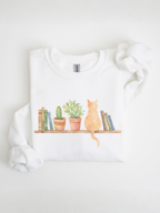 folded white sweatshirt with watercolor cat sitting among books and plants displayed on a clean background