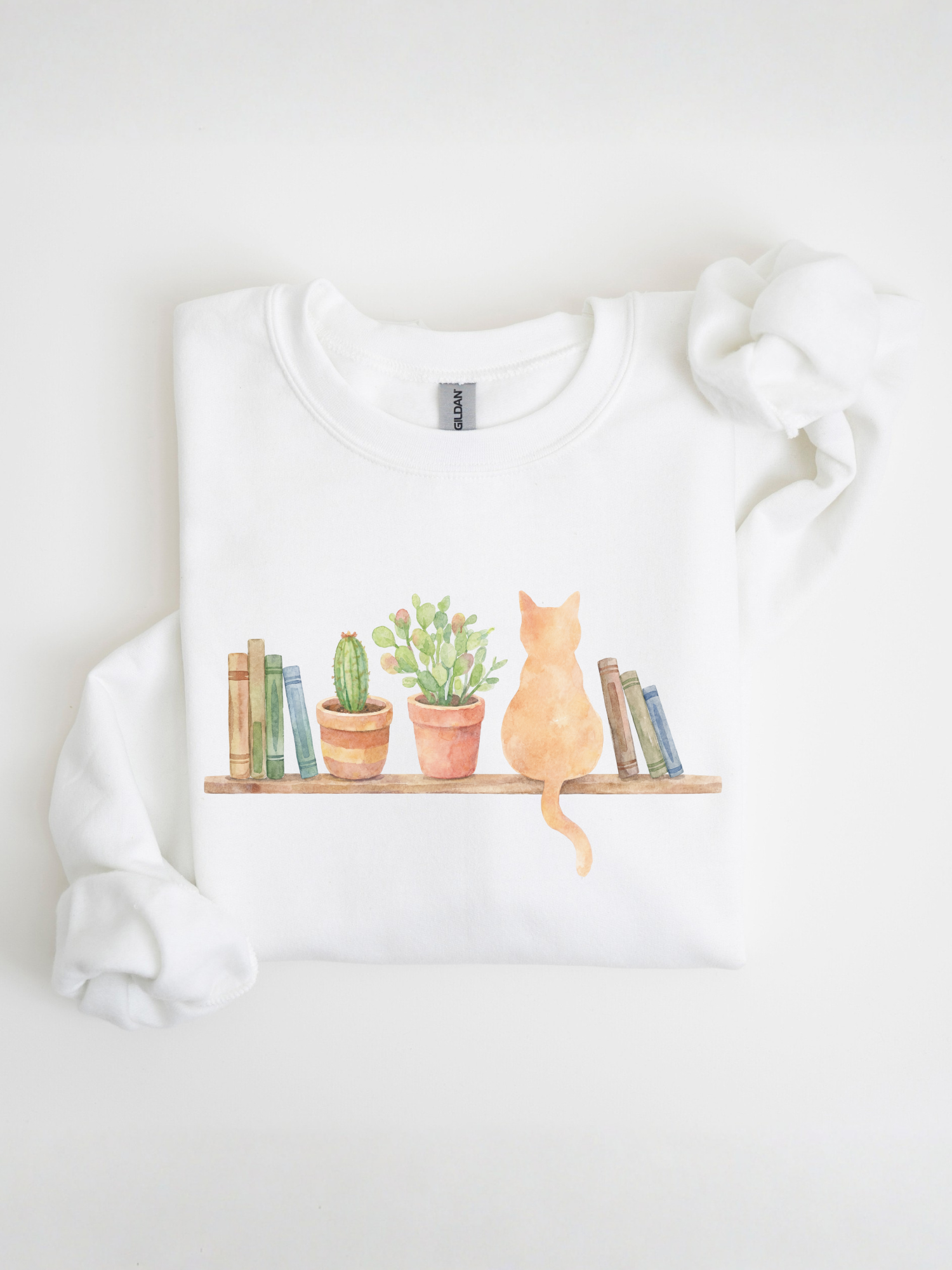 folded white sweatshirt with watercolor cat sitting among books and plants displayed on a clean background