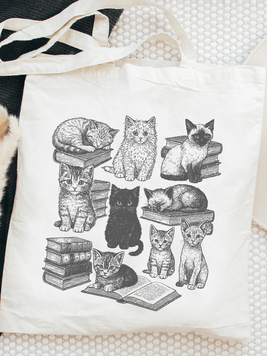 aesthetic bookish tote bag with illustrated cats lounging and reading on books, styled flat on a textured white background