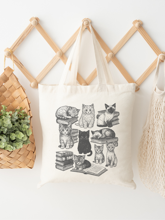 canvas tote bag with hand-drawn vintage cats and stacked books design hanging on wooden accordion wall rack beside woven basket