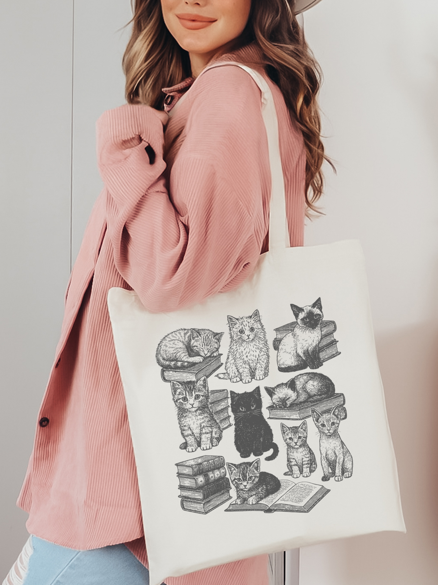 woman holding canvas tote bag with cute black and white cat and book print, styled with pink corduroy jacket and hat