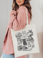 woman holding canvas tote bag with cute black and white cat and book print, styled with pink corduroy jacket and hat