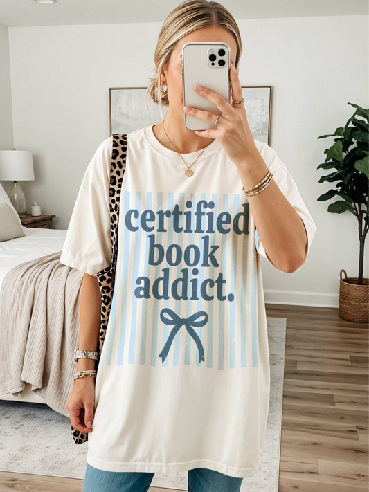 woman wearing certified book addict comfort colors t shirt with blue striped background and bow graphic in cozy bedroom mirror selfie
