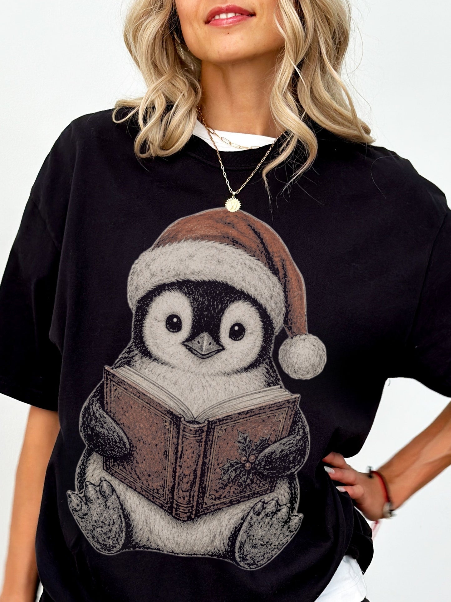 Model wearing a Black Comfort Colors bookish shirt with a cute penguin in a Santa hat reading a vintage Christmas book