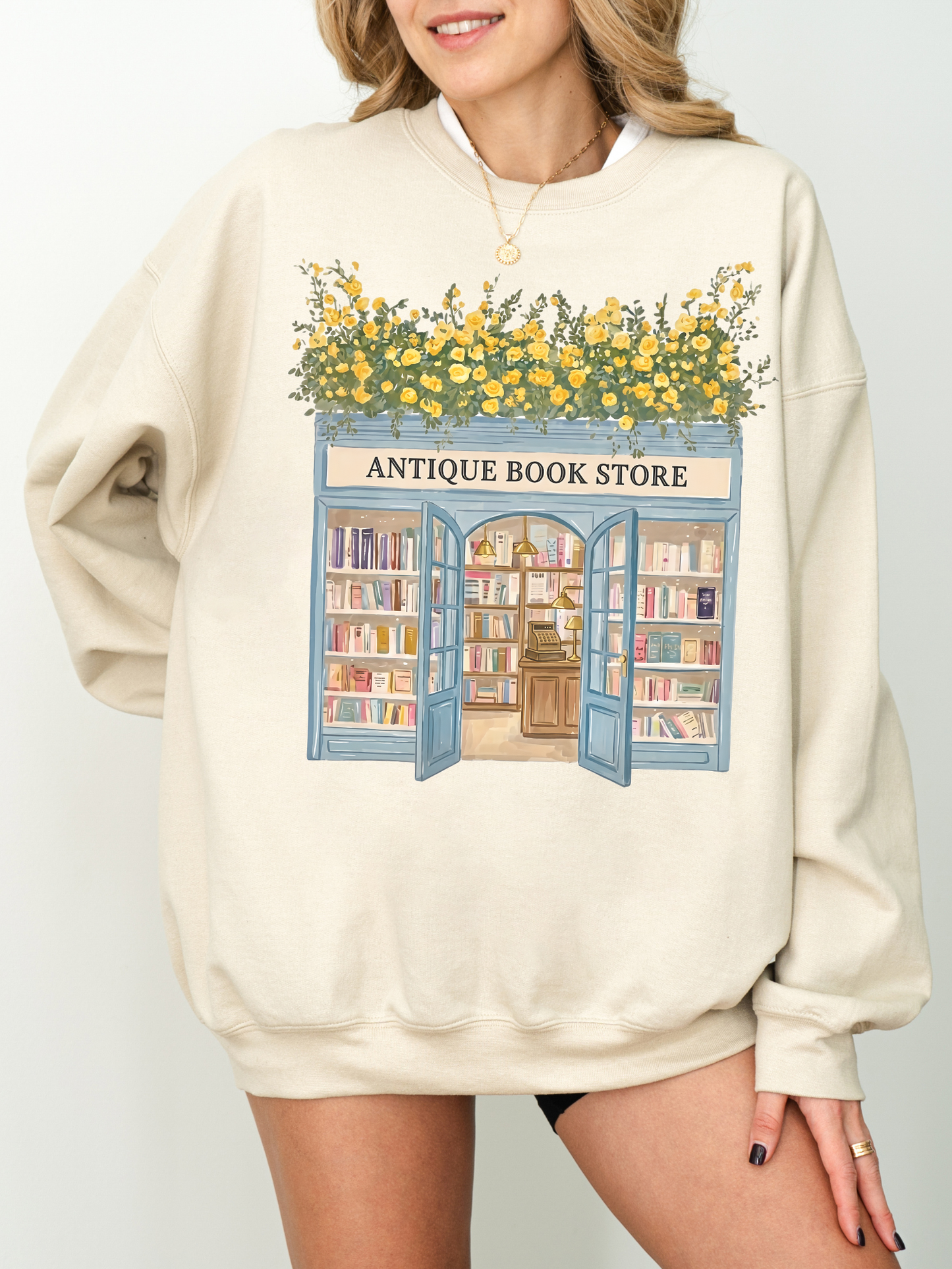 Woman wearing a sand sweatshirt with a vintage bookstore illustration featuring open doors, shelves of books, and yellow flowers styled in a soft neutral aesthetic
