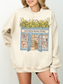 Woman wearing a sand sweatshirt with a vintage bookstore illustration featuring open doors, shelves of books, and yellow flowers styled in a soft neutral aesthetic