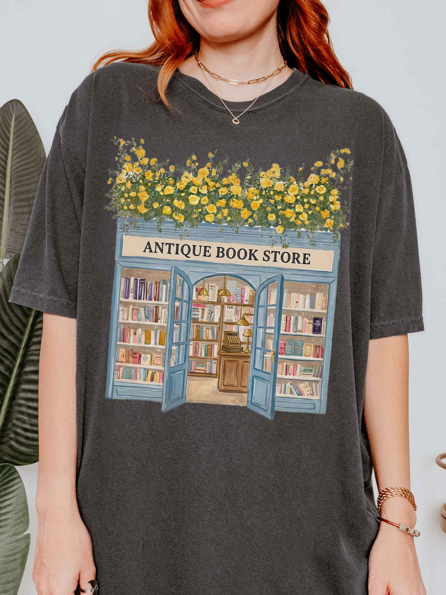 Woman wearing a pepper t shirt with a detailed antique bookstore design featuring open doors, bookshelves, and yellow flowers styled with minimal jewelry indoors