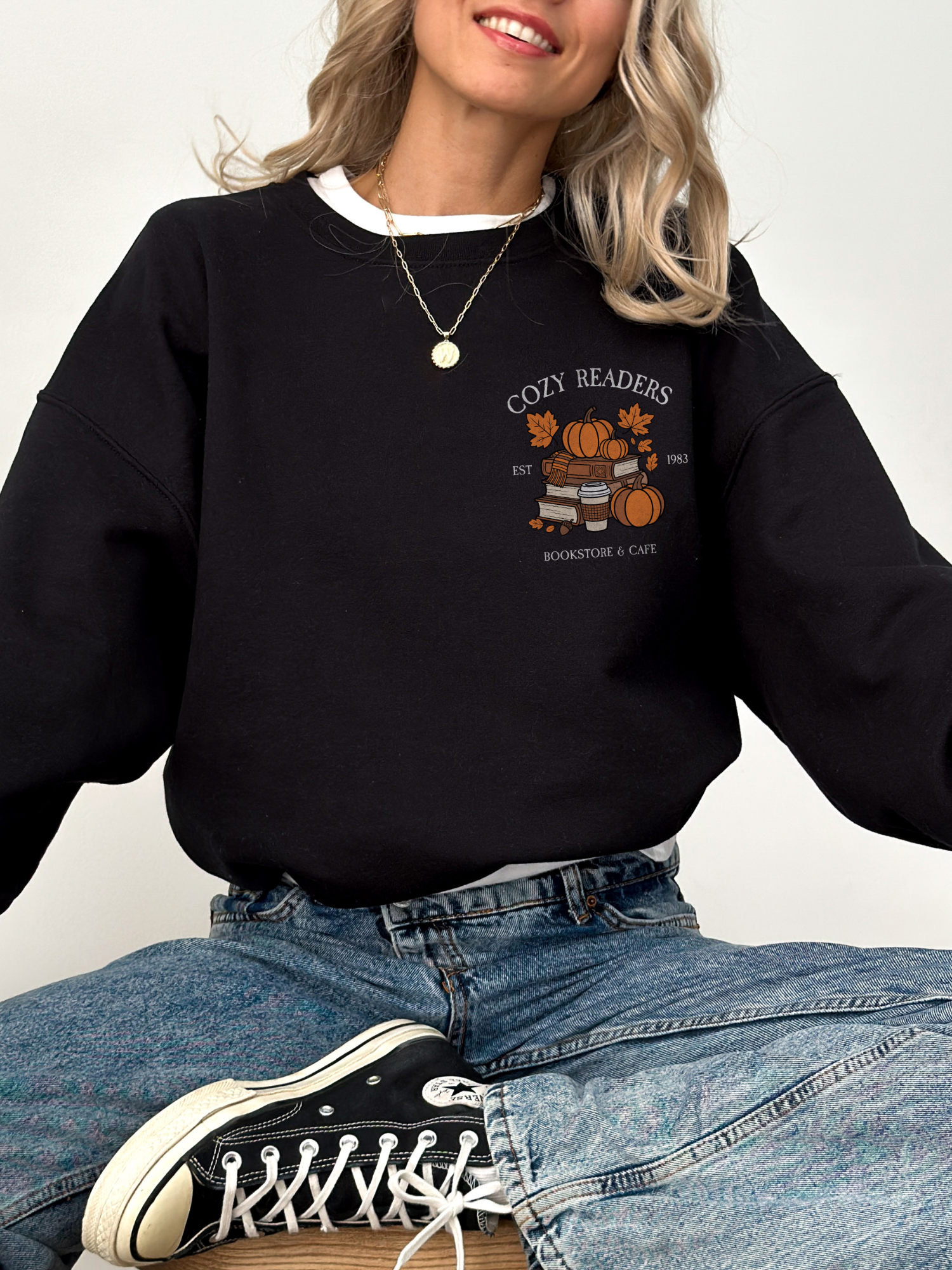 Woman seated in black Cozy Readers sweatshirt featuring a small front chest print with pumpkins, books, and coffee design. Styled with classic jeans and sneakers.