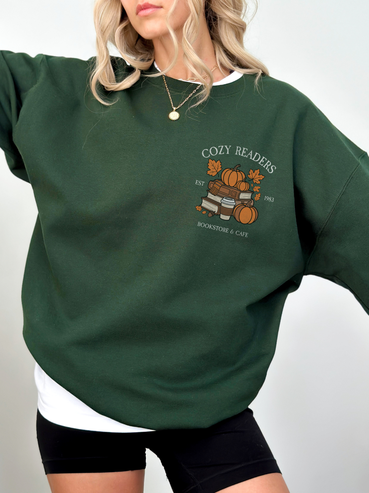 Woman wearing forest green Cozy Readers sweatshirt with vintage fall graphic on chest. Features stacked books, pumpkin, takeaway coffee, and falling leaves.