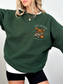 Woman wearing forest green Cozy Readers sweatshirt with vintage fall graphic on chest. Features stacked books, pumpkin, takeaway coffee, and falling leaves.