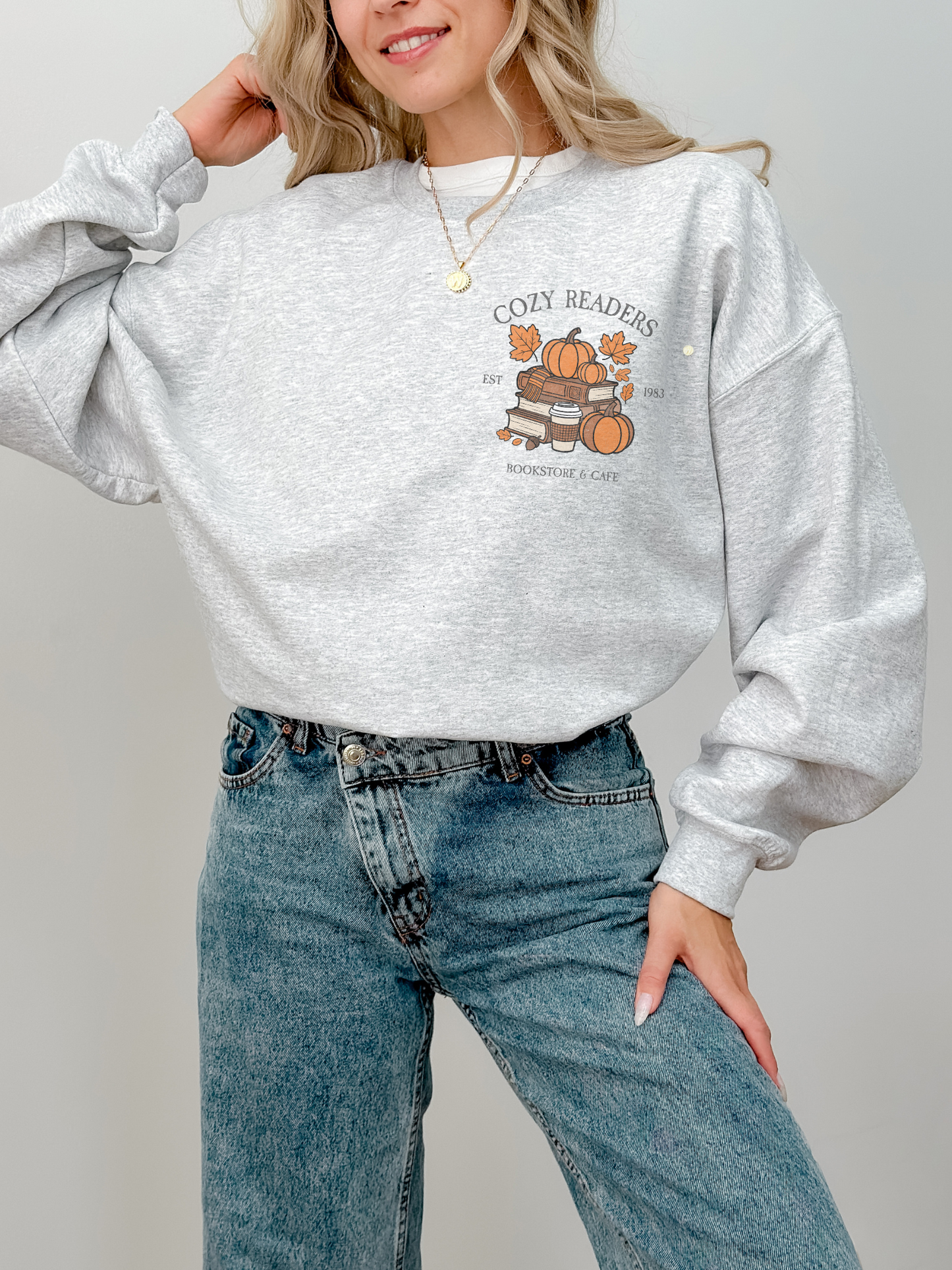 Woman wearing light grey Cozy Readers sweatshirt featuring a small front chest graphic of pumpkins, books, coffee cup, and leaves with text Cozy Readers Bookstore and Cafe EST 1983. Styled with blue denim jeans and gold necklace.