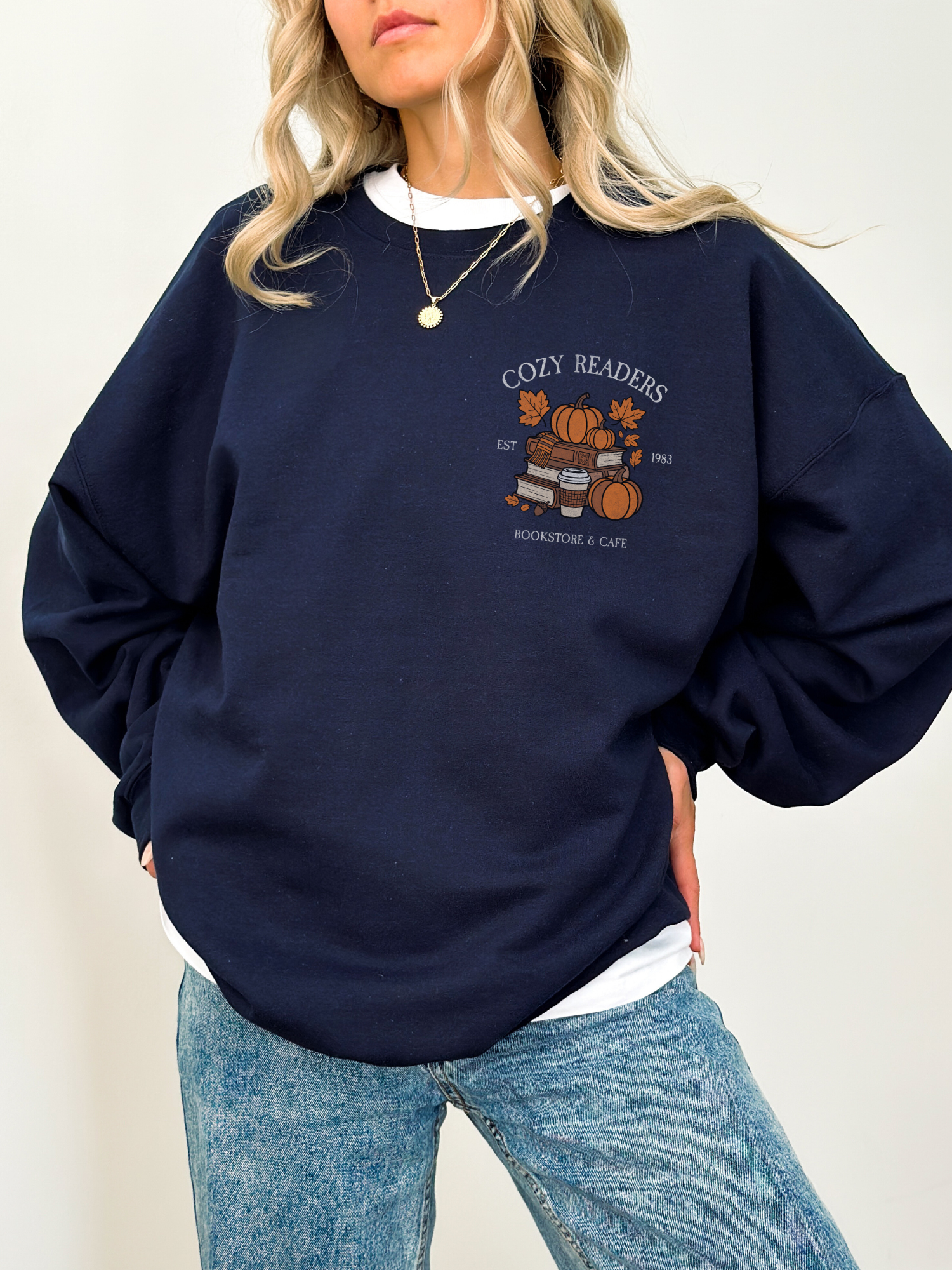 Woman wearing forest green Cozy Readers sweatshirt with vintage fall graphic on chest. Features stacked books, pumpkin, takeaway coffee, and falling leaves.