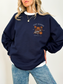Woman wearing forest green Cozy Readers sweatshirt with vintage fall graphic on chest. Features stacked books, pumpkin, takeaway coffee, and falling leaves.