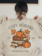 Back view of oversized sand Gildan sweatshirt featuring Cozy Readers Bookstore and Cafe graphic with pumpkins, books, and acorns. Model has hair in a bun and is pulling at her collar.